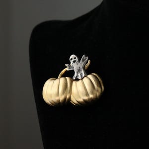 May include: A Halloween-themed brooch featuring a silver ghost figure atop two gold-colored pumpkins. The brooch is attached to a black surface, creating a striking contrast. This festive accessory is perfect for Halloween.