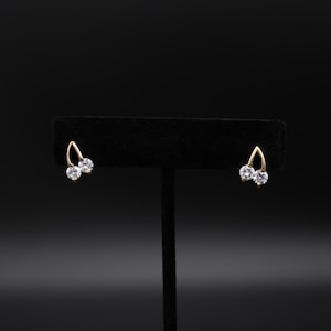 May include: A pair of gold-colored stud earrings, each featuring two clear, round-cut gemstones. The earrings are displayed on a black velvet jewelry stand against a black background. The earrings have a curved, open design.