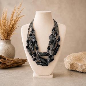 Puede incluir: Un collar de múltiples hilos que presenta una combinación de cuentas negras lisas, cuentas esféricas de color gris mate y componentes texturizados alargados y retorcidos de color carbón, ensartados en hilos de alambre metálico oscuro.