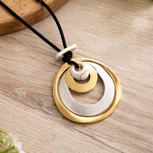 Peut inclure: Un collier pendentif avec un cordon noir. Le pendentif présente trois cercles concentriques en or et argent, avec un centre blanc marbré. Le collier est posé sur une surface en bois.