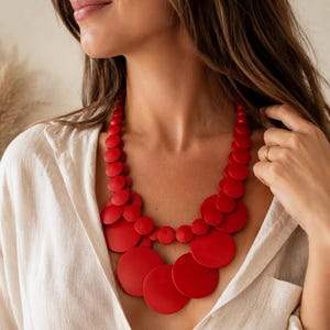Può includere: Una collana importante a più strati caratterizzata da numerose perle in legno rosso, lisce e circolari, di varie dimensioni. Le perle sono disposte a cascata, creando un accessorio audace e vivace che poggia sulla zona della clavicola.