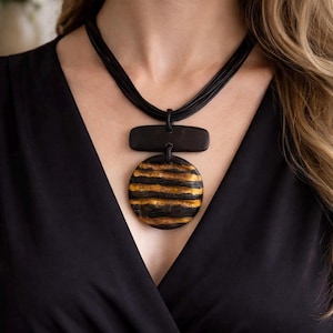 Könnte beinhalten: Eine schwarze Halskette mit einem kreisförmigen Anhänger mit horizontalen Gold- und Schwarzstreifen. Die Halskette hat einen rechteckigen schwarzen Akzent über dem Anhänger und mehrere schwarze Kordeln.