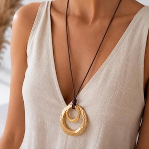 Op de afbeelding: Een ketting met een donkerbruin leren koord en een hanger bestaande uit twee in elkaar grijpende goudkleurige cirkelvormige ringen met een gehamerde, getextureerde afwerking. De hanger hangt aan het koord, dat bovenaan is vastgeknoopt.