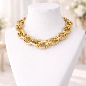 Könnte beinhalten: Eine goldfarbene Halskette mit einem klobigen Kettendesign. Die Halskette wird auf einer weißen Mannequin-Büste präsentiert. Die Kettenglieder haben ein strukturiertes, gewebtes Muster, und der Gesamtstil ist kühn. Der Hintergrund ist leicht verschwommen.