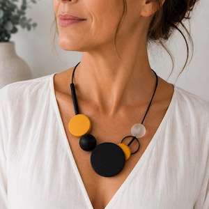 Op de afbeelding: Een statement ketting met een dun zwart koord versierd met diverse geometrische vormen, waaronder een grote matzwarte cirkel, een kleinere mosterdgele cirkel, een massieve zwarte bol en een doorschijnende, matwitte kraal.