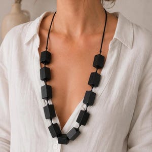 Puede incluir: Un collar largo con diez cuentas rectangulares de color negro mate ensartadas en un cordón negro fino. Las cuentas están dispuestas en una secuencia vertical, colgando del cuello en forma de V, contrastando con una camisa blanca lisa.