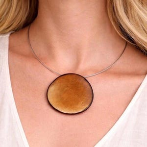 Op de afbeelding: Een ketting met een grote, ronde, barnsteenkleurige hanger met een glad, gepolijst oppervlak. De hanger hangt aan een dunne, zilverkleurige metalen draad die om de hals rust, wat zorgt voor een strakke en eenvoudige uitstraling.