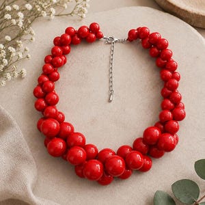 Peut inclure: Un collier composé de multiples grappes de perles sphériques rouge vif et brillantes de tailles variées. Le collier est doté d'une chaînette d'extension en métal argenté avec un fermoir mousqueton, permettant d'ajuster la longueur autour du cou.