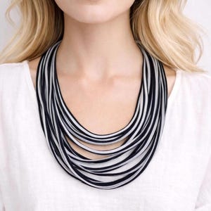 Puede incluir: Un collar llamativo de múltiples hilos con cordones finos que alternan entre negro y gris claro. El collar consta de varios bucles circulares superpuestos que caen alrededor del cuello, creando un patrón texturizado sobre una blusa blanca.