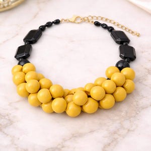 Puede incluir: Un collar llamativo con una combinación de cuentas negras y amarillas. Cuenta con una cadena y cierre dorados. El diseño incluye grandes cuentas negras geométricas y cuentas amarillas redondas más pequeñas, creando un contraste audaz.