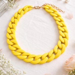 Puede incluir: Un collar de cadena amarillo brillante. Presenta eslabones grandes e interconectados y un cierre dorado. Esta llamativa pieza se exhibe sobre una superficie blanca, con detalles florales en el fondo. Un accesorio de moda.
