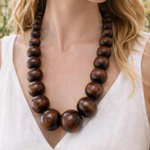 Può includere: Una collana di perline di legno marrone scuro. La collana presenta grandi perline rotonde che si riducono gradualmente di dimensione. Le perline hanno una finitura lucida e la collana è indossata sopra un top bianco.