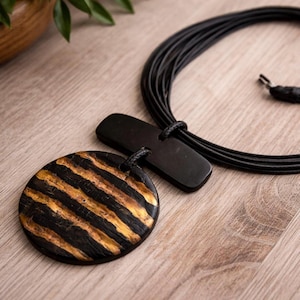 Puede incluir: Un collar con colgante a rayas negras y doradas con un cordón de cuero negro a juego. El colgante es redondo con un patrón de rayas. El collar también incluye un cierre rectangular negro.
