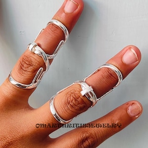 以下が含まれることがあります： 指にフィットするようにデザインされた、複雑で絡み合ったデザインの銀の指輪3つ。指輪は手の親指と中指につけられています。