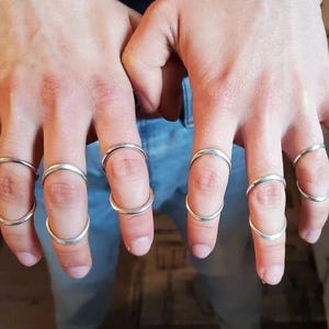 May include: A person's hands wearing multiple silver rings on each finger. The rings are simple and minimalist, with a rounded shape.