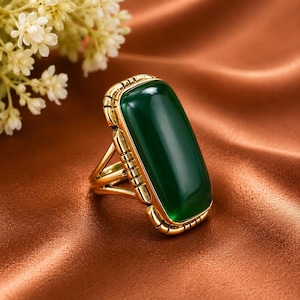 Pode incluir: Um anel de tom dourado com uma pedra grande, retangular e lisa de cor verde-escura, engastada numa armação de metal com detalhes decorativos ranhurados nas laterais. A banda consiste em vários fios metálicos finos e entrecruzados.