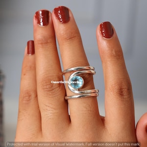 May include: A silver ring with a light blue gemstone. The ring has a unique design with a spiral shape around the gemstone. The ring is on a person's finger. The text "Omarthriti jewelry" is visible.
