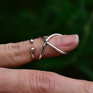 May include: A silver ring with a criss-cross design and two small balls on the side. The ring is worn on a finger.