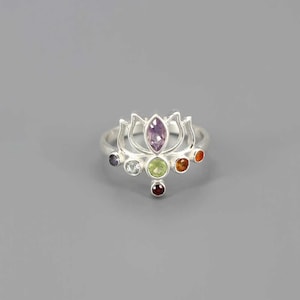 Puede incluir: Un anillo de plata con un diseño de flor de loto, con una gema púrpura central y seis gemas de colores más pequeñas. El diseño del anillo incorpora varios colores, incluyendo púrpura, verde, naranja y rojo, sobre una banda plateada.