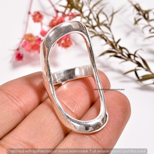 May include: A silver ring with an unusual elongated oval design. The ring has a rectangular band connecting to the oval shape. The jewellery is held in a hand against a soft background.