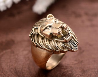 May include: A gold-toned ring with a detailed lion head design. The lion's mane is intricately sculpted and the ring has a wide band.