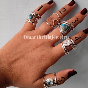 Könnte beinhalten: Eine Hand zeigt fünf Silberringe, jeder mit einem einzigartigen Design. Die Ringe weisen verschiedene Edelsteine auf, darunter Türkis und ein blauer Stein. Der Text "Omarthritisjewelry" ist sichtbar.