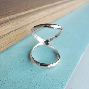 May include: A silver-colored ring with an infinity symbol design. The ring is made of a shiny metal and has a smooth, polished surface. The ring is displayed on a light blue surface.