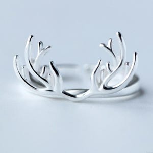 May include: A silver-colored ring featuring an antler design. The ring's antlers are detailed and symmetrical, with multiple points extending upwards. The ring is set against a light background, highlighting its metallic sheen. The ring is a piece of jewelry.
