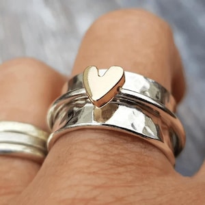 May include: A silver ring with a textured finish and a gold heart-shaped detail. The ring is on a finger, with another silver ring in the background. Jewellery.