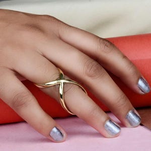 May include: A gold-toned ring with a criss-cross design. The ring is worn on the index finger of a hand with silver nail polish.
