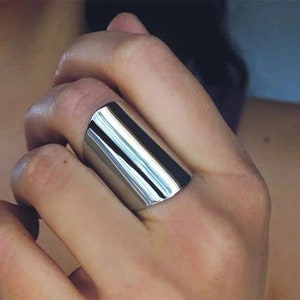 Pode incluir: Um anel de prata polida com um design retangular. O anel é liso e refletivo, captando a luz. A joia é usada em um dedo, mostrando seu estilo minimalista.