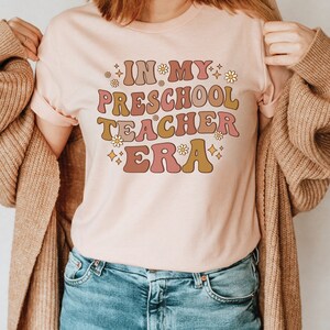 May include: A pink t-shirt with the text "In My Preschool Teacher Era" in a retro font with pink, yellow, and brown colors and small flower accents.