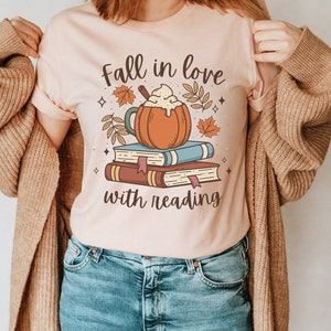 Pode incluir: Uma camiseta rosa com o texto "Fall in love with reading" e uma ilustra&ccedil;&atilde;o de desenho animado de um latte de especiarias de ab&oacute;bora em cima de uma pilha de livros.