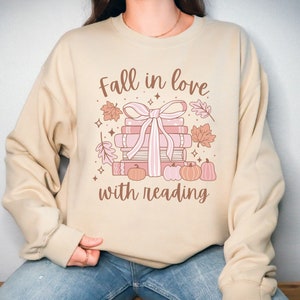 Pu&ograve; includere: Una felpa beige con il testo "Fall in love with reading" e una grafica di una pila di libri legati con un nastro rosa e circondati da foglie autunnali e zucche.