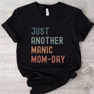 May include: Black t-shirt with the text "Just Another Manic Mom-Day" in a vintage style font with colourful letters.
