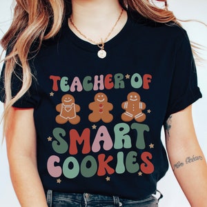 May include: A navy blue t-shirt with the text "Teacher of Smart Cookies" in colorful, retro-style lettering. Three gingerbread men are featured above the text, each with a different colored icing design.