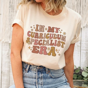 May include: A beige t-shirt with a retro-style graphic that reads "In My Curriculum Specialist Era" with pink, brown, and gold lettering and small flower accents.