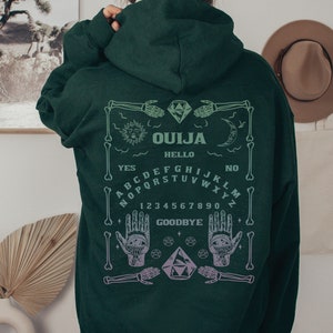 May include: Dark green hoodie with a Ouija board design on the back. The design includes the word "OUJIA", the alphabet, numbers, and the words "YES", "NO", "HELLO", and "GOODBYE". The design also features a sun, moon, hands, and diamonds.
