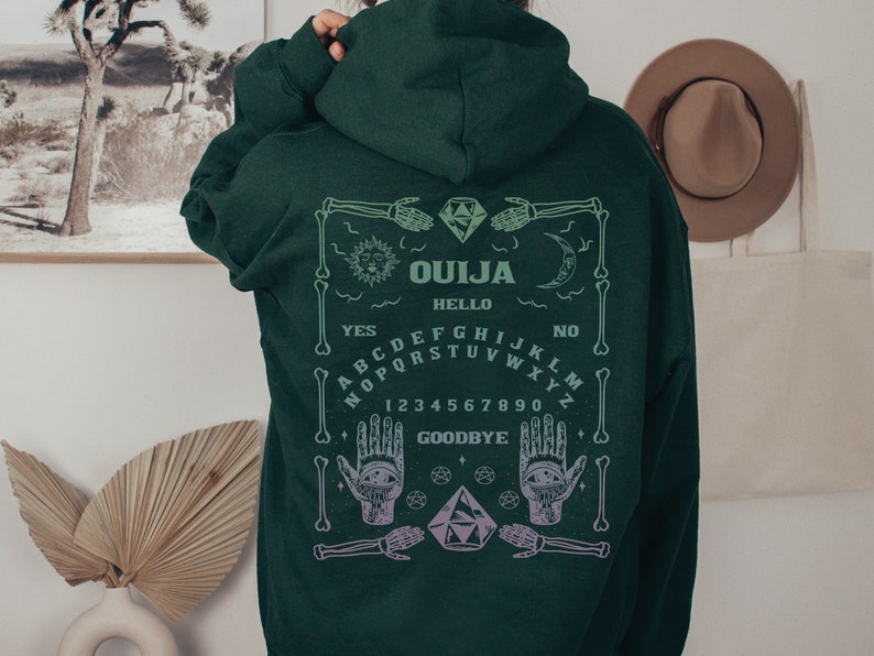 May include: Dark green hoodie with a Ouija board design on the back. The design includes the word "OUJIA", the alphabet, numbers, and the words "YES", "NO", "HELLO", and "GOODBYE". The design also features a sun, moon, hands, and diamonds.