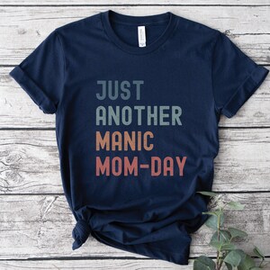 May include: A navy blue t-shirt with the text "JUST ANOTHER MANIC MOM-DAY" in a vintage style font with a gradient of colours.