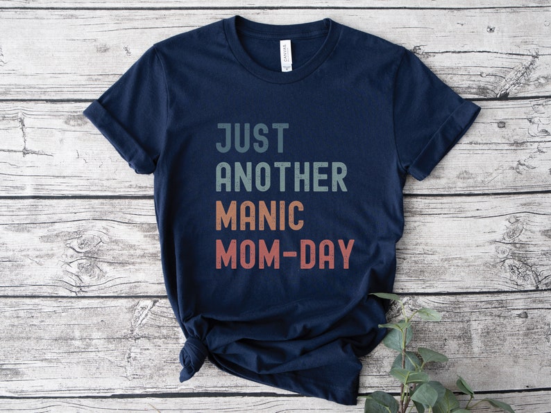 May include: A navy blue t-shirt with the text "JUST ANOTHER MANIC MOM-DAY" in a vintage style font with a gradient of colours.