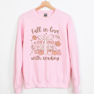 Pu&ograve; includere: Felpa rosa con un design grafico di una pila di libri legati con un nastro e zucche. Il testo "Fall in love with reading" &egrave; stampato sulla felpa.