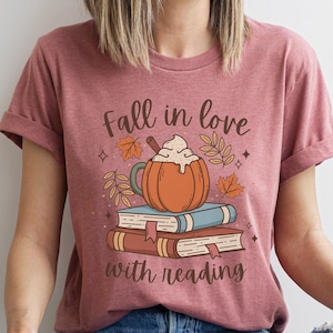 Pode incluir: Uma camiseta rosa com o texto "Fall in love with reading" e um gr&aacute;fico de um latte de especiarias de ab&oacute;bora com um pau de canela e uma pilha de livros.