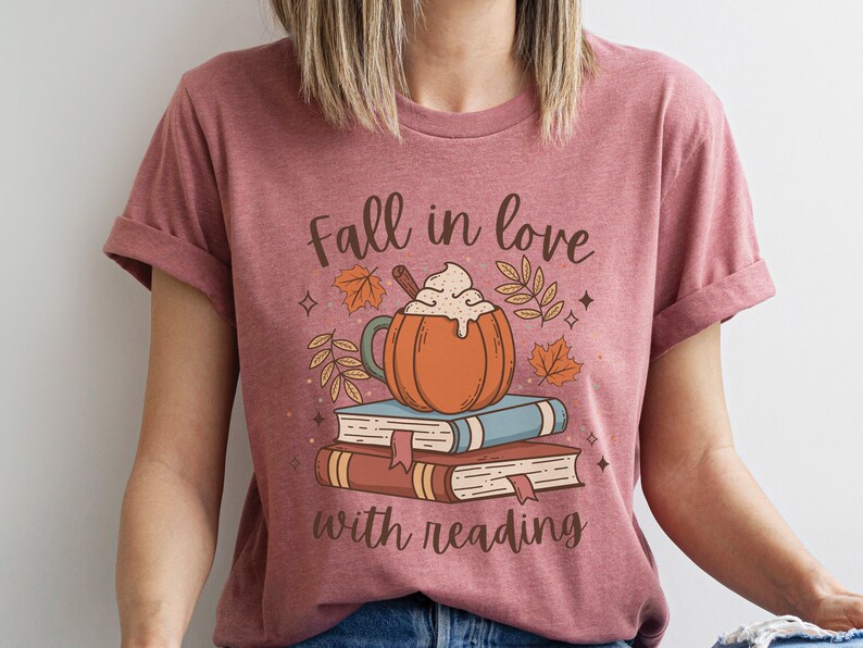 Pode incluir: Uma camiseta rosa com o texto "Fall in love with reading" e um gr&aacute;fico de um latte de especiarias de ab&oacute;bora com um pau de canela e uma pilha de livros.