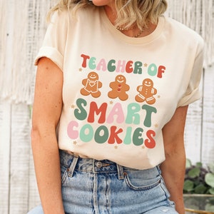 May include: A beige t-shirt with the text "Teacher of Smart Cookies" in colorful, retro-style lettering. Three gingerbread cookie illustrations are above the text.