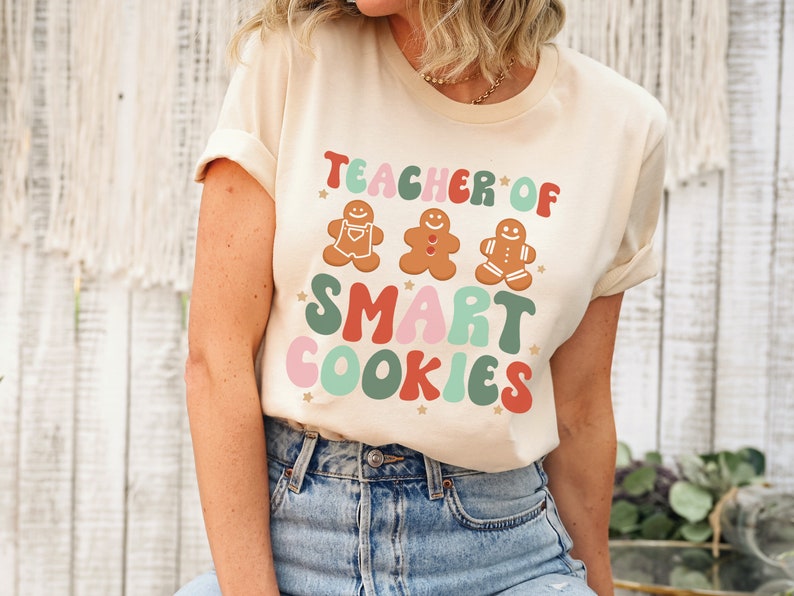 May include: A beige t-shirt with the text "Teacher of Smart Cookies" in colorful, retro-style lettering. Three gingerbread cookie illustrations are above the text.