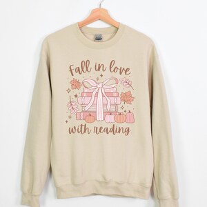 Pu&ograve; includere: Una felpa beige con la scritta "Fall in love with reading" stampata sul davanti. Il testo &egrave; in lettere marroni e rosa. C'&egrave; una pila di libri con un nastro rosa legato intorno e alcune foglie autunnali e zucche.