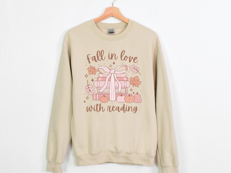 Pu&ograve; includere: Una felpa beige con la scritta "Fall in love with reading" stampata sul davanti. Il testo &egrave; in lettere marroni e rosa. C'&egrave; una pila di libri con un nastro rosa legato intorno e alcune foglie autunnali e zucche.