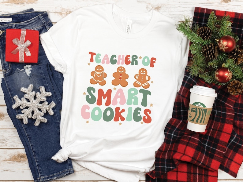 May include: White t-shirt with a festive design featuring gingerbread men and the text "Teacher of Smart Cookies".