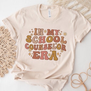 May include: A beige t-shirt with the text "In My School Counselor Era" in a retro font with pink, orange, and yellow colors. The text is surrounded by small white flowers and gold glitter.